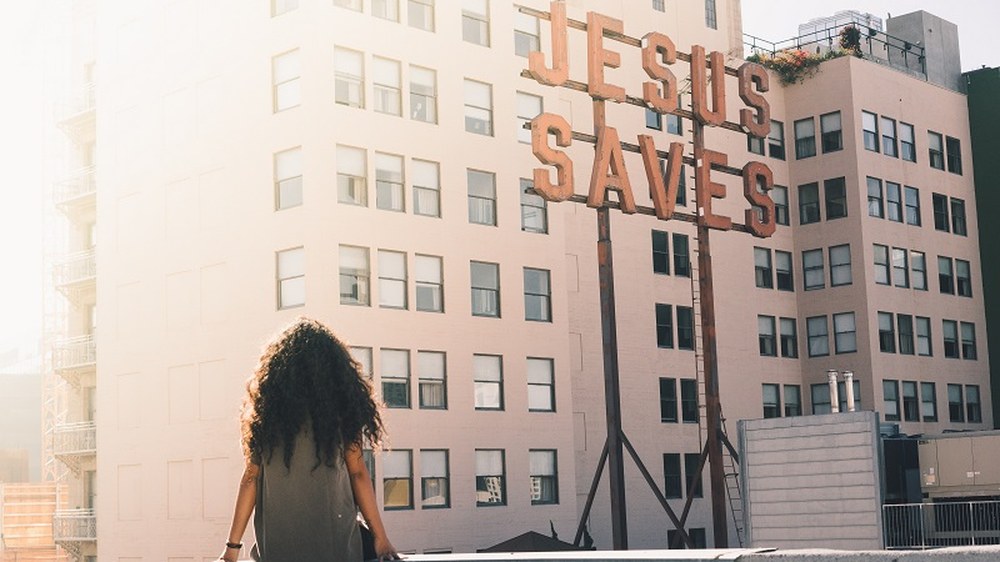 Woman on a rooftop looking toward a Jesus Saves sign at golden hour